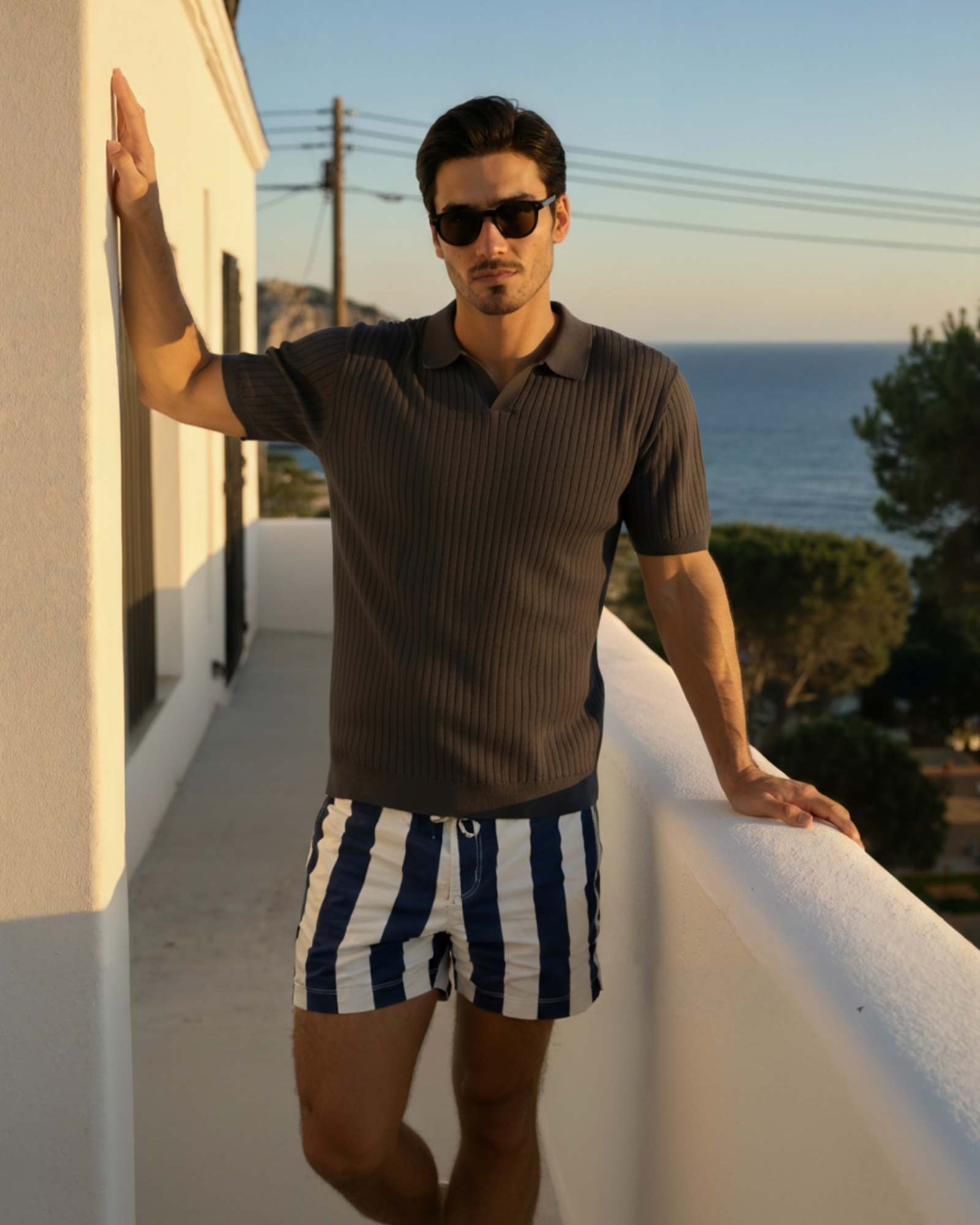 Man wearing sunglasses and a brown ribbed polo shirt with striped shorts standing on a balcony with ocean view.